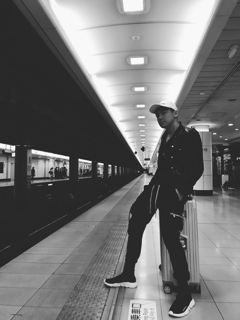 Black-and-white photo of a traveler in a cap leaning on a suitcase on a long, brightly lit train platform