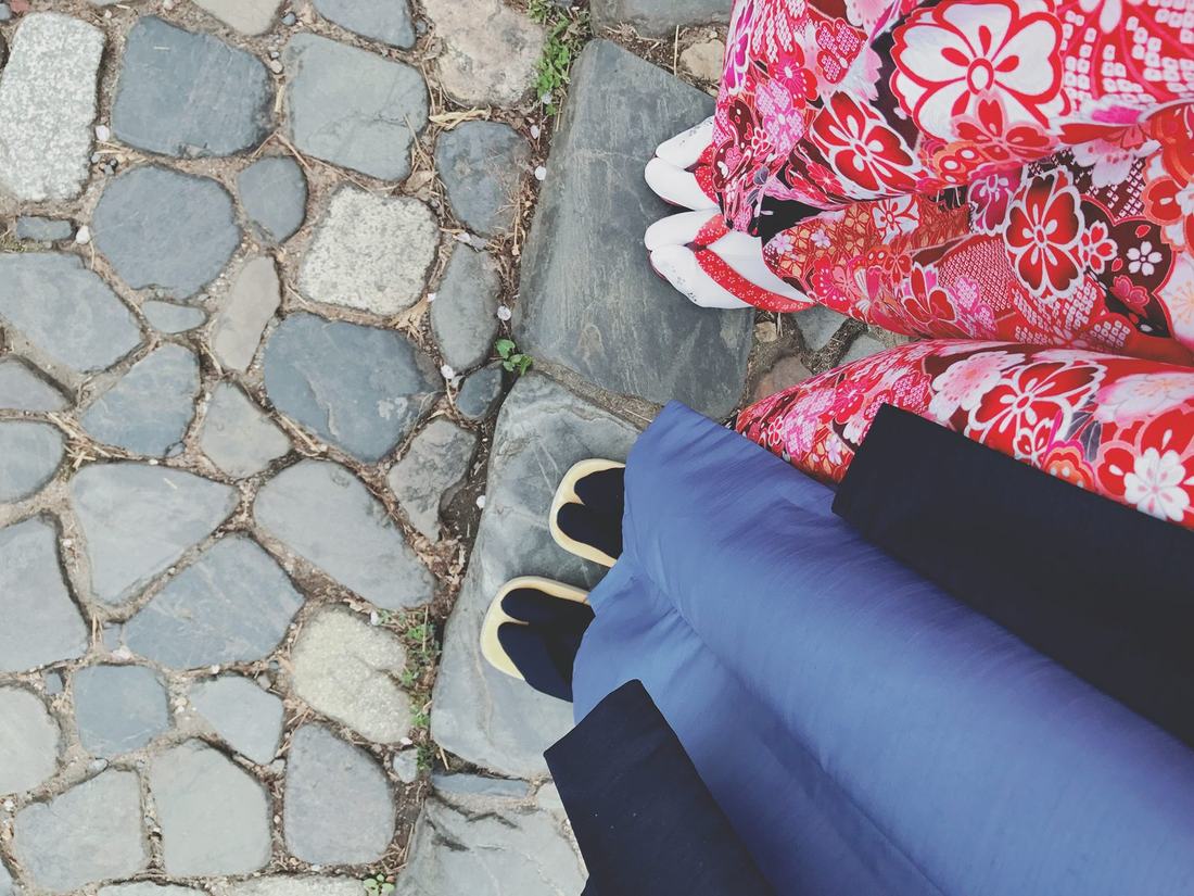 Looking down at two pairs of feet in sandals and kimono, one red and floral, one blue, on old paving stones