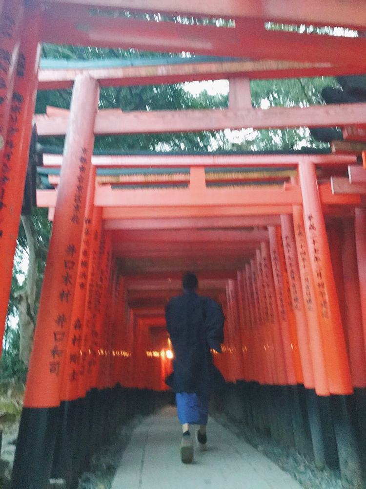 A figure in a dark kimono walking away through a tunnel of vermilion torii gates at dusk